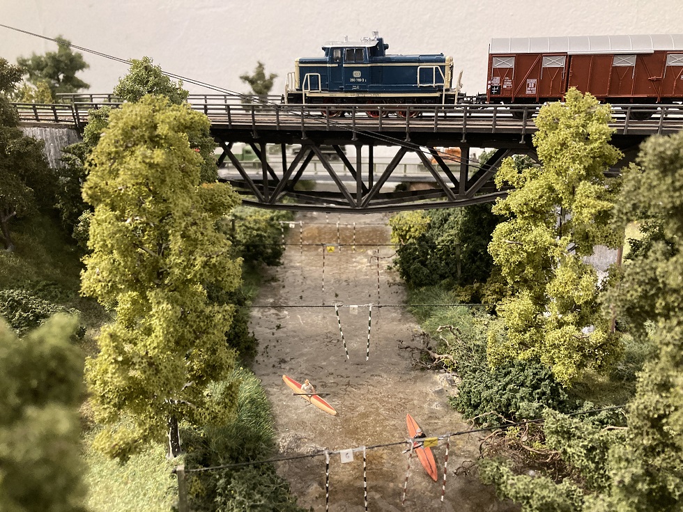 Modellzug mit Diesellok auf der Stahlbrücke über die Rednitz, darunter Bäume und das Flussufer
